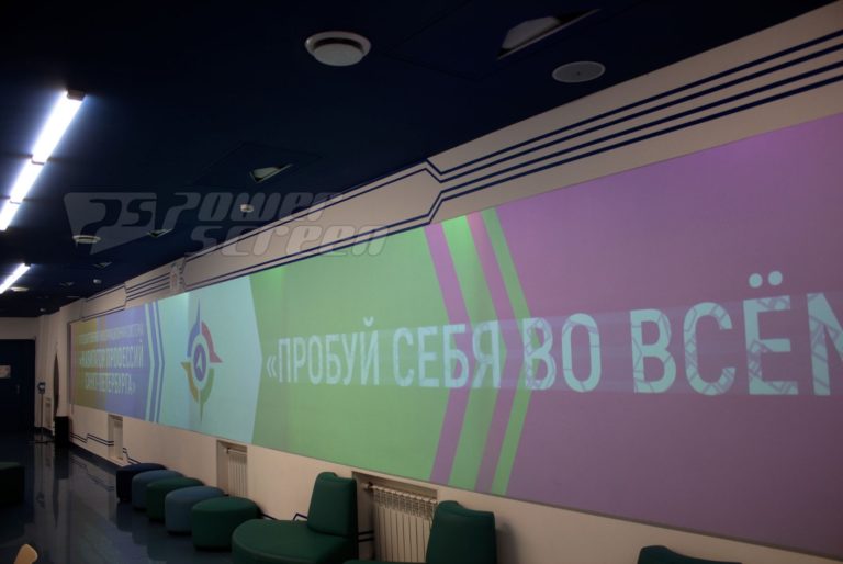 Длинная изогнутая стена имеет проекцию с русским текстом в ярких цветах. В комнате современное освещение, синие мягкие сиденья и гладкий дизайн.