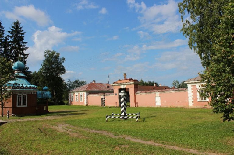 Травянистый двор с деревянной часовней с синими куполами и крестом слева. Длинное розовое здание с красной крышей тянется по всему фону. Посередине двора стоит полосатый черно-белый столб. Деревья и голубое небо окружают сцену.